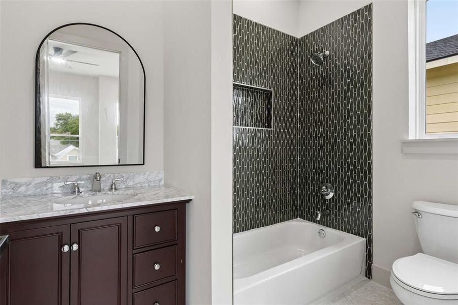 Bathroom featuring a dark wood vanity with a light stone countertop, an arched mirror, an undermount sink with a chrome faucet, a white bathtub with dark geometric wall tiling, and a built-in shower niche. Bathroom featuring a dark wood vanity with a light stone countertop, an arched mirror, an undermount sink with a chrome faucet, a white bathtub with dark geometric wall tiling, and a built-in shower niche.