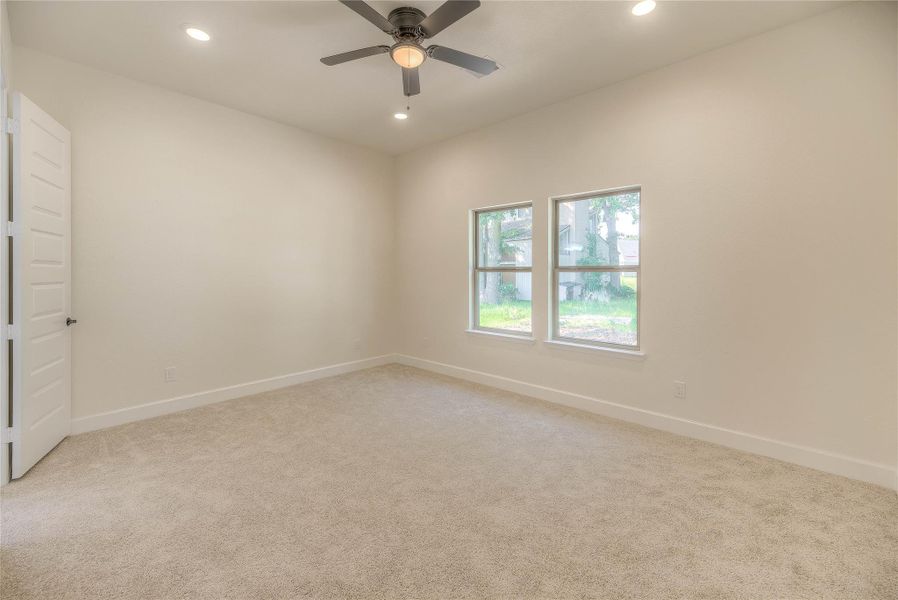 This photo shows a bright, spacious room with neutral-colored walls and carpet. It features a ceiling fan, recessed lighting, and two large windows offering a view outside.