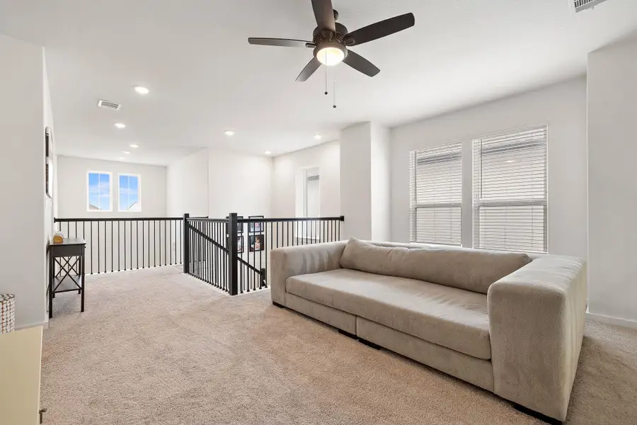 Carpeted loft area featuring a ceiling fan with integrated lighting, recessed lighting, multiple windows with blinds, and iron balusters