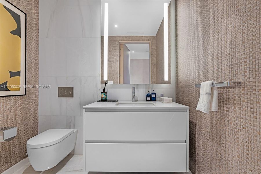 Floating vanity with a smooth white finish and countertop basin, wall-mounted commode, integrated vertical LED mirror lighting, and textured wall tile