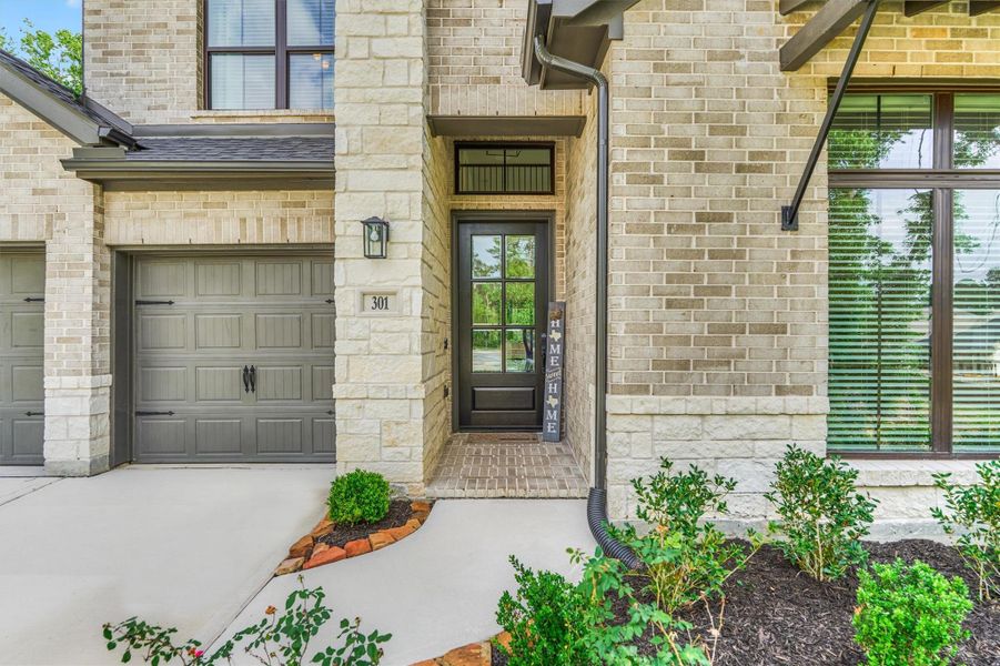 This photo showcases a charming brick home exterior with a welcoming front entrance. It features a modern garage door, a pathway with landscaped greenery, and a stylish front door under a sheltered porch.