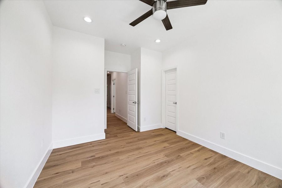 Another angle of the bedroom highlights its comfortable proportions and clean contemporary styling. Warm wood-look flooring runs throughout, while recessed lighting and a modern ceiling fan add function and visual appeal. Neutral walls create a versatile backdrop for furnishings, and nearby doors provide convenient access to the hallway and closet space.