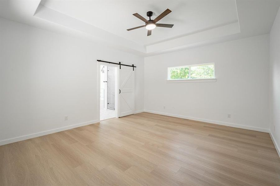 Unfurnished bedroom with a tray ceiling, a barn door, light wood finished floors, and ceiling fan Unfurnished bedroom with a tray ceiling, a barn door, light wood finished floors, and ceiling fan