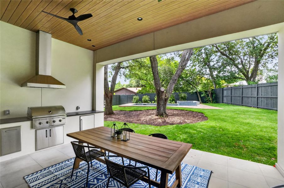 An inviting outdoor kitchen and dining area flows seamlessly into a lush backyard with mature trees and a modern pool.