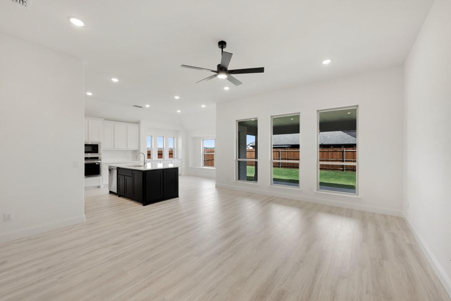 Open living area with light wood floors, ceiling fan, and kitchen with dark island visible in background