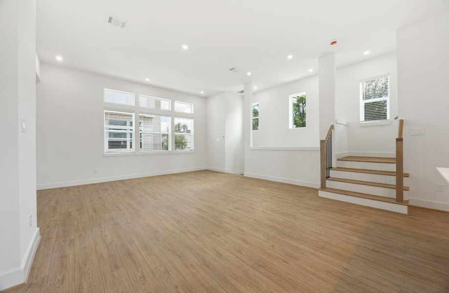Expansive main living area with high ceilings, recessed lighting, warm wood-look flooring, and a wall of windows that brings in beautiful natural light. The open layout provides generous flexibility for living and dining arrangements, while the stairway adds architectural interest and an easy connection to the home’s upper-level spaces.