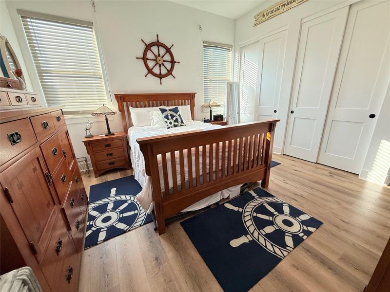 This bedroom features a nautical theme with elegant wood furniture, a ship wheel wall decor, and matching navy rugs. It has large windows for ample natural light, a comfortable bed, and spacious closets. Perfect for a serene and stylish retreat.