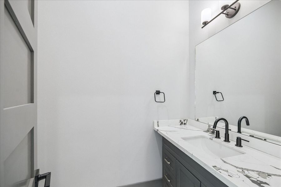 Quartz counters with striking veining and matte black fixtures bring a modern touch to the powder bath. A spacious mirror and wall-mounted lighting ensure a bright, inviting atmosphere that balances practicality with style.