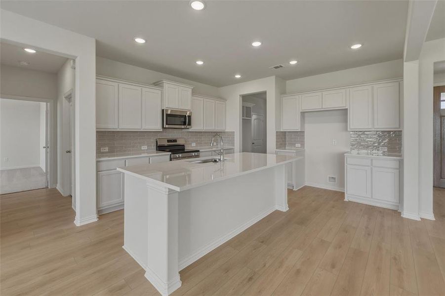 Kitchen featuring a large central island, white cabinetry, stainless steel appliances, tile backsplash, and light wood-finish flooring Kitchen featuring a large central island, white cabinetry, stainless steel appliances, tile backsplash, and light wood-finish flooring