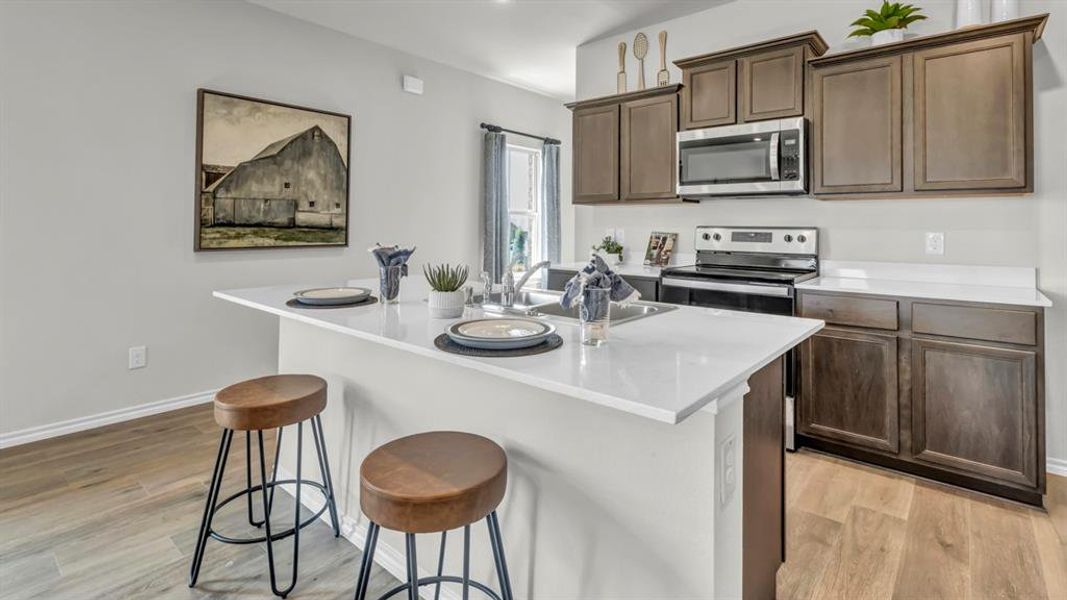 Kitchen featuring a white countertop island with an undermount sink, warm wood-finish flooring, dark wood cabinetry, a stainless steel microwave, and a black electric range Kitchen featuring a white countertop island with an undermount sink, warm wood-finish flooring, dark wood cabinetry, a stainless steel microwave, and a black electric range