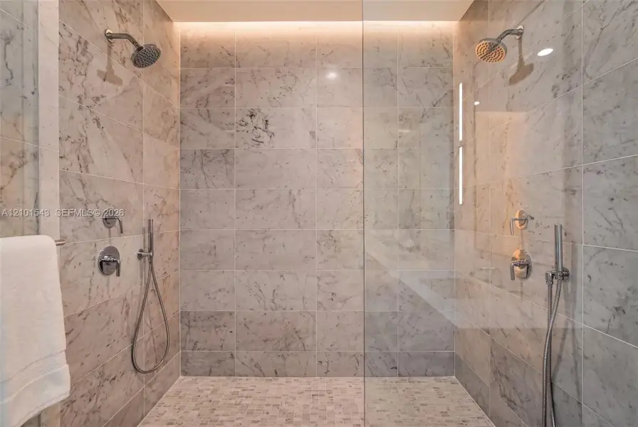 Walk-in shower featuring large-format stone wall tiling, mosaic stone flooring, dual shower heads with handheld sprayers, a frameless glass enclosure, and recessed overhead accent lighting