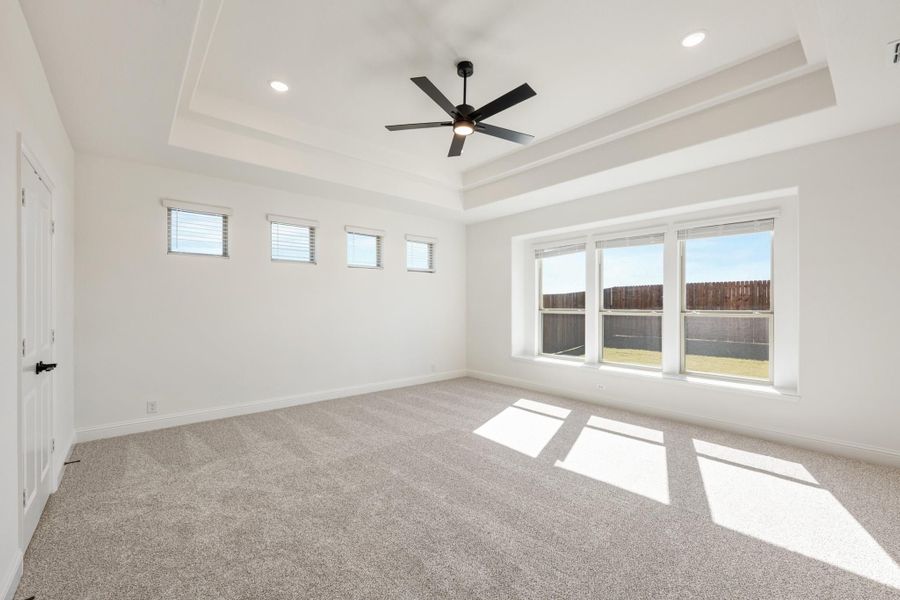 Empty bedroom with tray ceiling, ceiling fan, carpet flooring, and large windows overlooking backyard Empty bedroom with tray ceiling, ceiling fan, carpet flooring, and large windows overlooking backyard