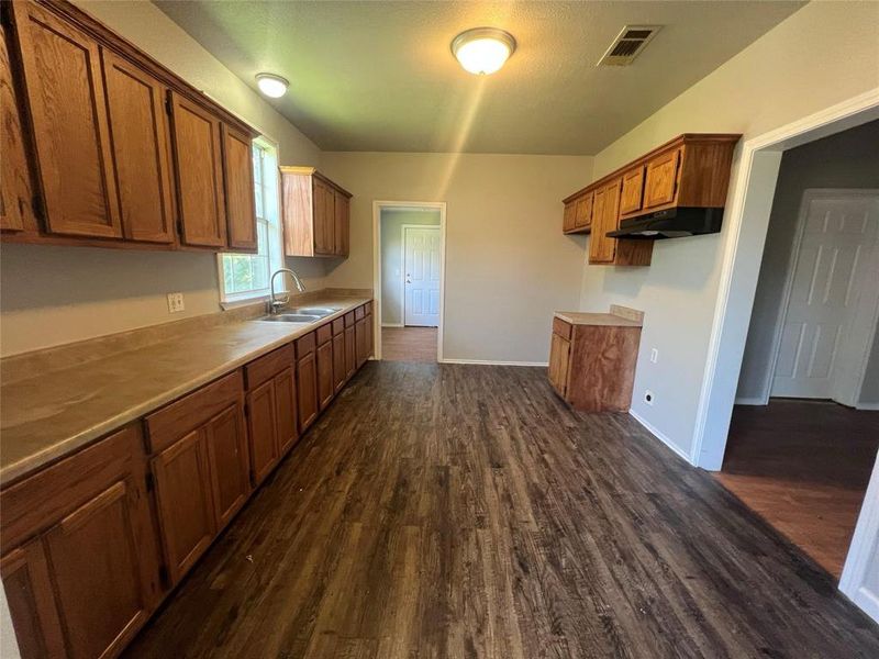 Kitchen featuring brown cabinets, dark wood-type flooring, light countertops, and under cabinet range hood Kitchen featuring brown cabinets, dark wood-type flooring, light countertops, and under cabinet range hood