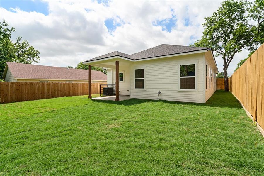 Expansive grassy yard with a covered patio featuring wooden support posts, a dark shingle roof, white siding, and a privacy fence