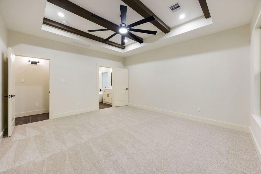 This spacious primary bedroom room features a modern design with a tray ceiling and exposed beams, a stylish ceiling fan, and large windows offering scenic views of greenery. The neutral carpet and wall colors create a warm, inviting atmosphere.