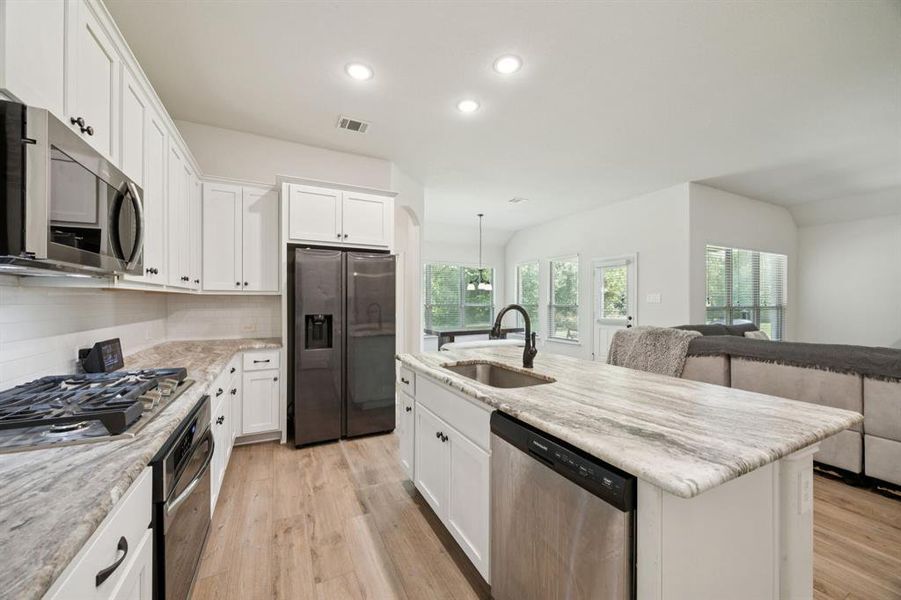 Open concept kitchen featuring white cabinets, granite counter tops, stainless steel appliances, recessed lighting, vinyl plank floors floors, and an island with sink.