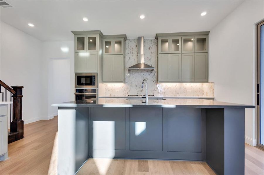 Kitchen featuring wall chimney range hood, light hardwood / wood-style flooring, built in microwave, decorative backsplash, and oven