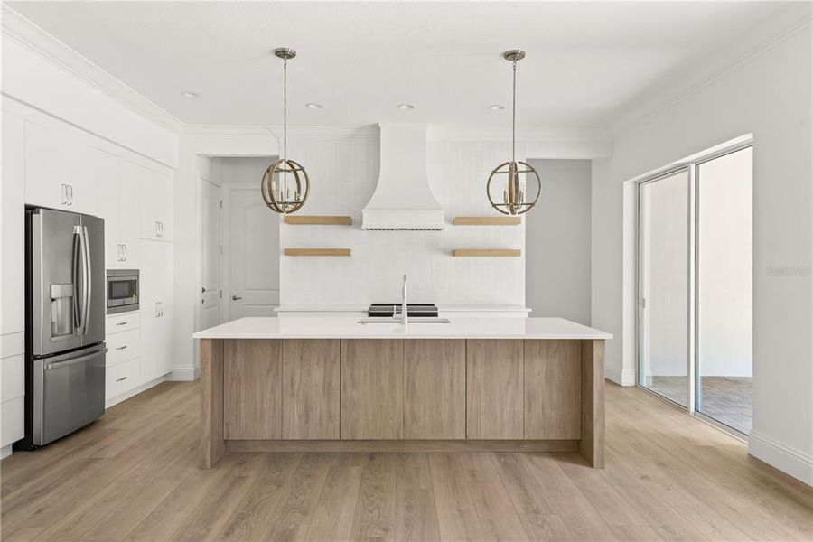 Modern kitchen featuring a center island with a white countertop and light wood cabinetry, stainless steel appliances, pendant lighting, and access to the lanai through sliding glass doors.