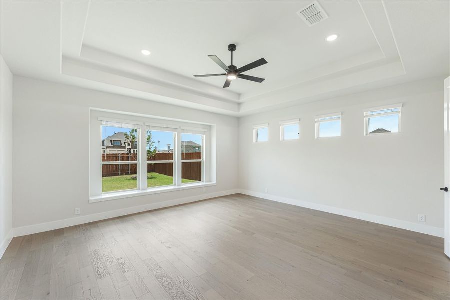 Empty bedroom with tray ceiling, ceiling fan, hardwood floors, and large windows overlooking backyard Empty bedroom with tray ceiling, ceiling fan, hardwood floors, and large windows overlooking backyard
