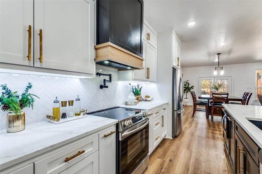 Kitchen with appliances with stainless steel finishes, white cabinets, light stone countertops, hanging light fixtures, and custom exhaust hood