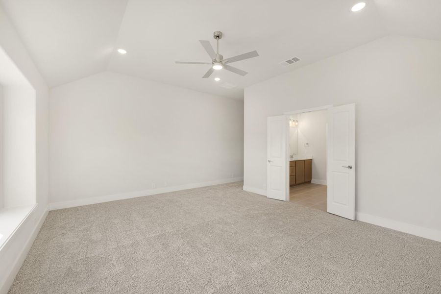 Empty bedroom with vaulted ceiling, ceiling fan, carpet flooring, and ensuite bathroom doorway visible. Empty bedroom with vaulted ceiling, ceiling fan, carpet flooring, and ensuite bathroom doorway visible.