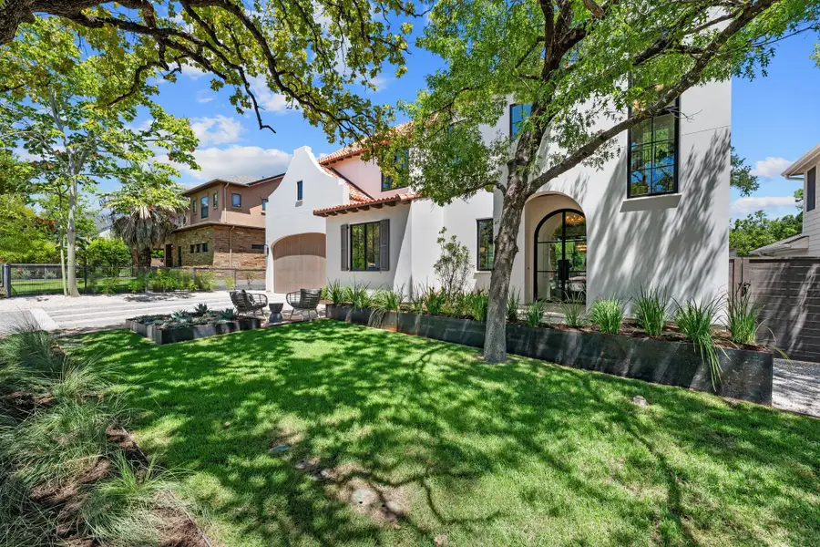 Mature trees shade a manicured lawn leading to the arched entryway of this stucco home.