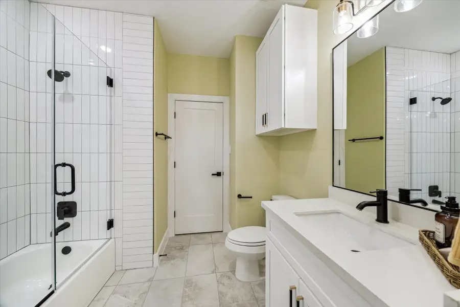 This bathroom features a modern design with a sleek glass-enclosed shower, white subway tile that is specially designed from floor to ceiling, and contemporary black fixtures. It includes a spacious vanity with a white countertop, ample storage even overhead for towels, and stylish lighting. The neutral color palette adds to the clean and fresh look.