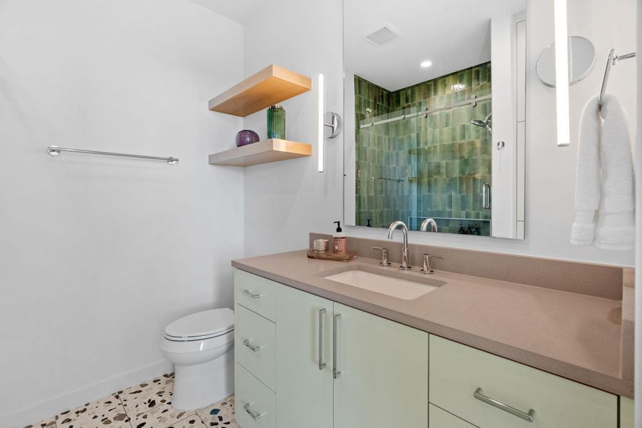 Bathroom featuring a light green vanity with integrated sink, a large mirror with vertical sconces, a shower with green subway tile, floating wood shelving, and patterned terrazzo-style flooring