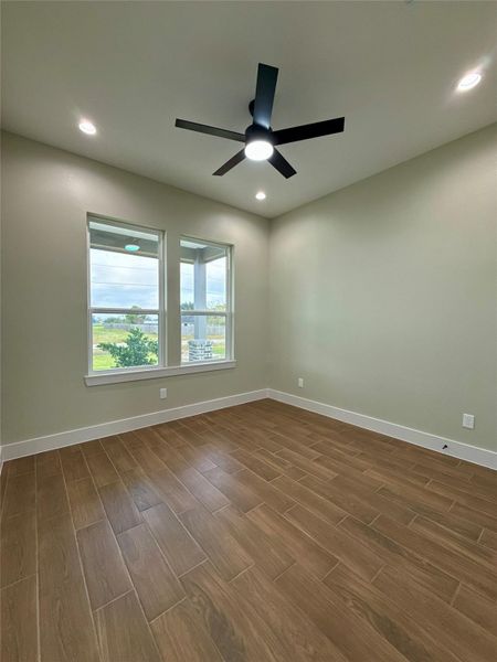This room features wood-look tile flooring, neutral walls, recessed lighting, and a modern ceiling fan. Large windows offer natural light and a view of the outdoors.