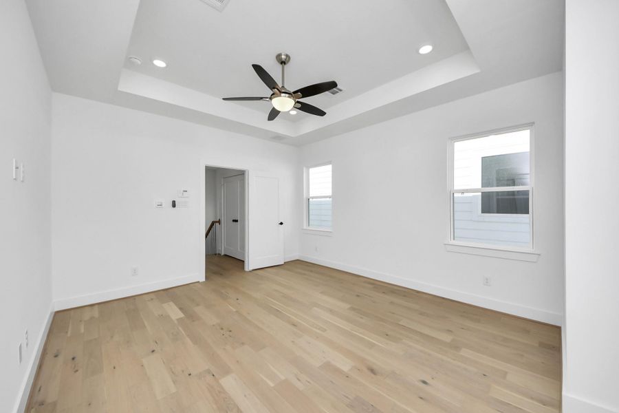 Bright, spacious room with light hardwood floors and white walls. Features a tray ceiling with recessed lighting and a ceiling fan. Two windows provide natural light, and there's an open doorway leading to another area.