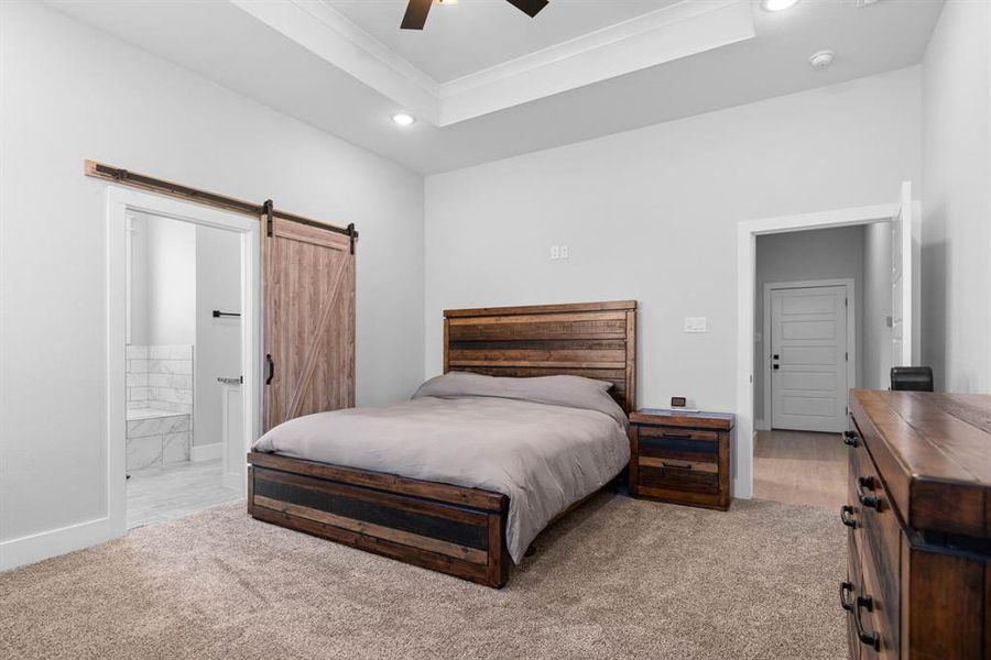 Carpeted bedroom featuring ceiling fan, ensuite bath, a barn door, and a tray ceiling