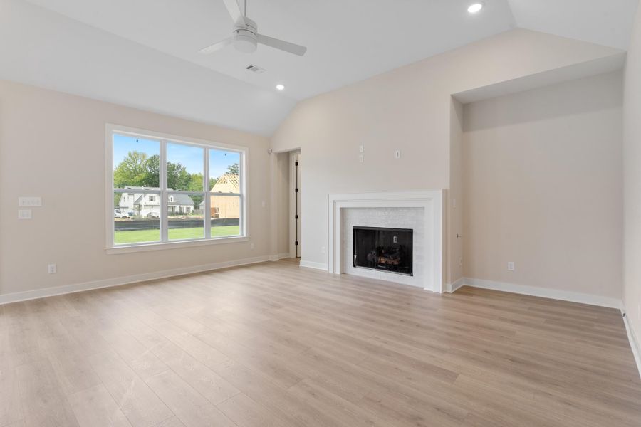 Vaulted ceiling with recessed lighting and ceiling fan, large three-pane window, wood-finish flooring, and a fireplace with a white mantel and light-toned tile surround Vaulted ceiling with recessed lighting and ceiling fan, large three-pane window, wood-finish flooring, and a fireplace with a white mantel and light-toned tile surround