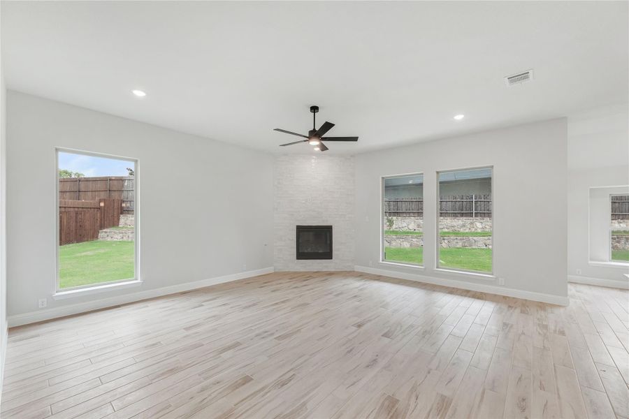 Open living room with light wood floors, brick fireplace, ceiling fan, and large windows overlooking backyard Open living room with light wood floors, brick fireplace, ceiling fan, and large windows overlooking backyard