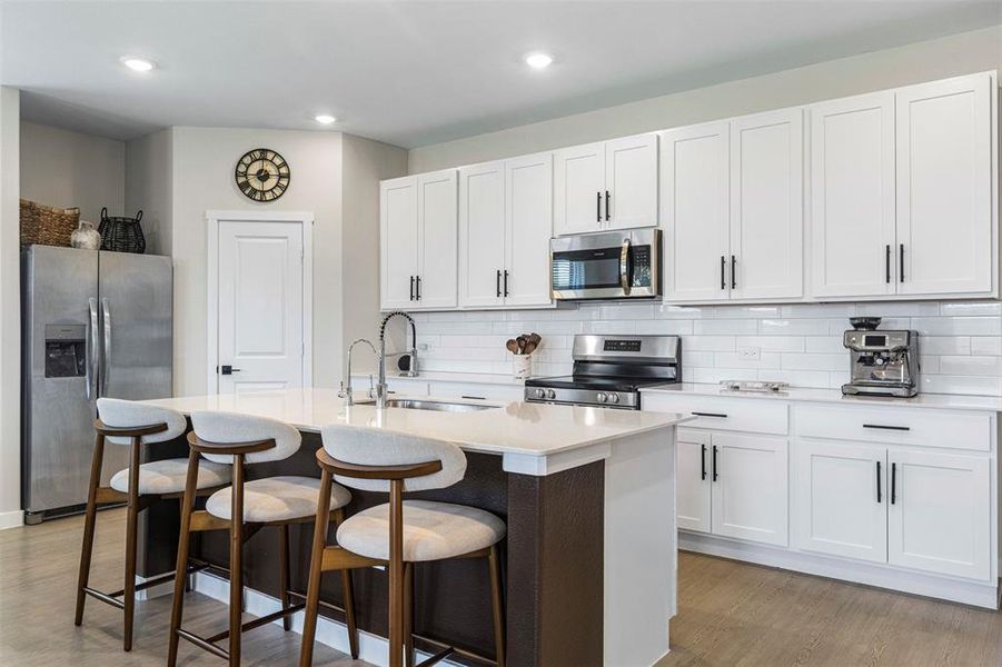 Modern kitchen featuring a central island with countertop seating, white shaker-style cabinetry, stainless steel appliances, white subway tile backsplash, and wood-finish flooring Modern kitchen featuring a central island with countertop seating, white shaker-style cabinetry, stainless steel appliances, white subway tile backsplash, and wood-finish flooring