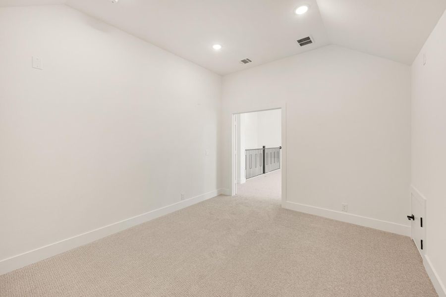 Empty bedroom with white walls, beige carpet, recessed lighting, and open door to hallway with stair railing visible
