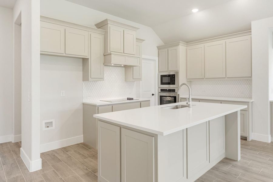Kitchen with gray cabinets, white quartz island with sink, tile backsplash, and built-in stainless steel ovens