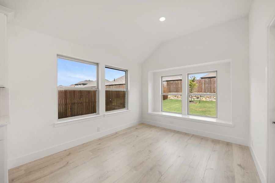 Empty room with vaulted ceiling, light wood floors, and four windows overlooking a fenced backyard
