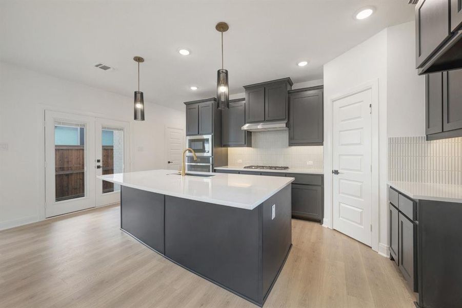 Kitchen featuring a central island with a white countertop and brass-finish faucet, dark gray cabinetry, integrated stainless steel appliances, and light wood-finish flooring