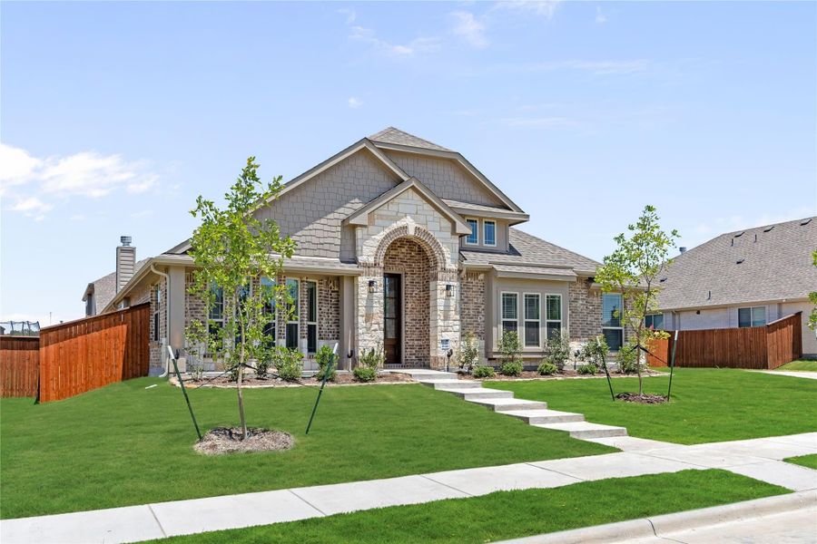 Single-story home with brick and stone exterior, arched entry, and manicured front lawn with young trees and concrete walkway.