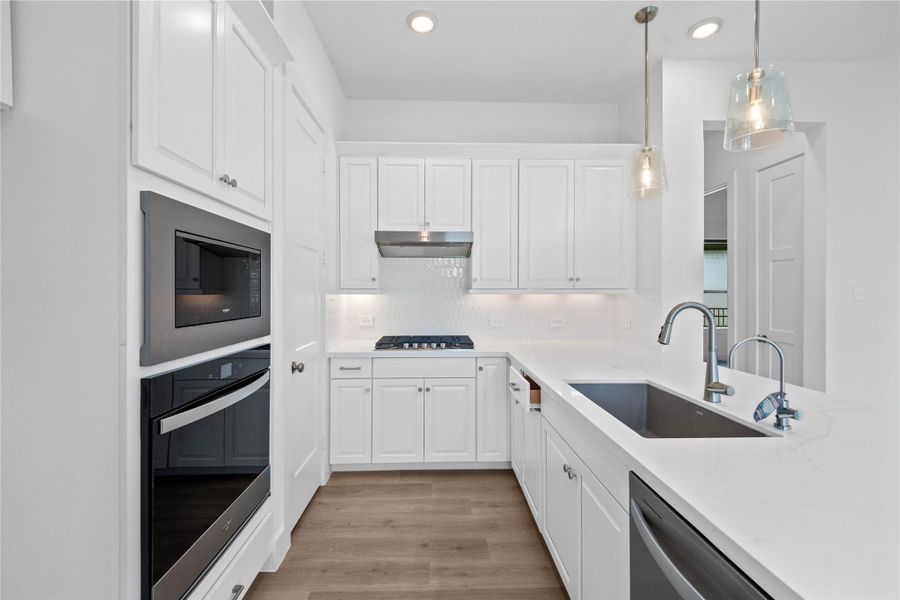 The open-concept gourmet kitchen is designed for both cooking and entertaining. It features elegant white cabinetry, pristine quartz countertops, and a large countertop that doubles as a functional breakfast bar. The cooking area is a chef's dream with a sleek stainless steel gas cooktop, hood vent, and oven/microwave wall unit, all complemented by a chic white tile backsplash.