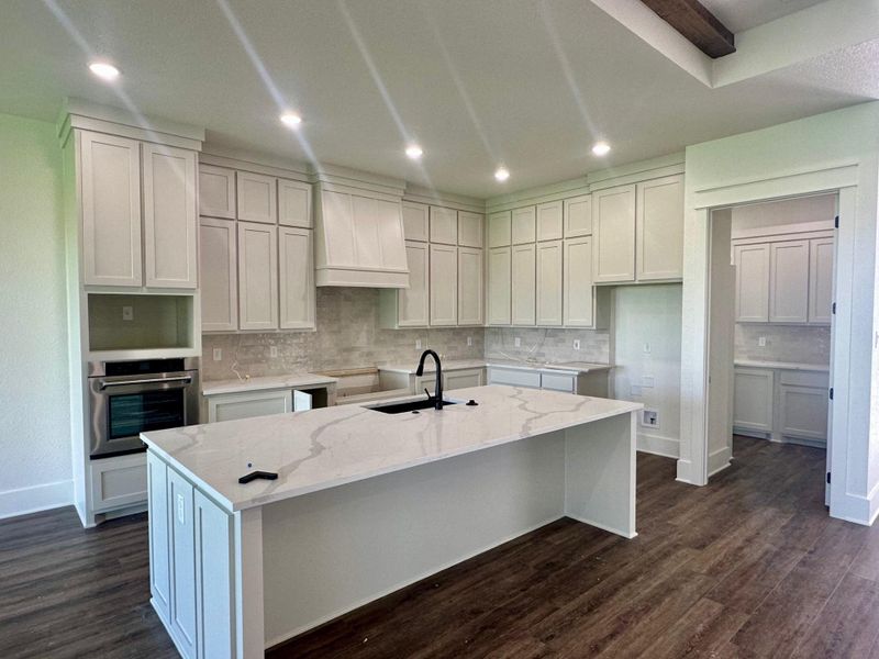 Kitchen with light stone counters, stainless steel oven, an island with sink, dark wood-type flooring, and decorative backsplash