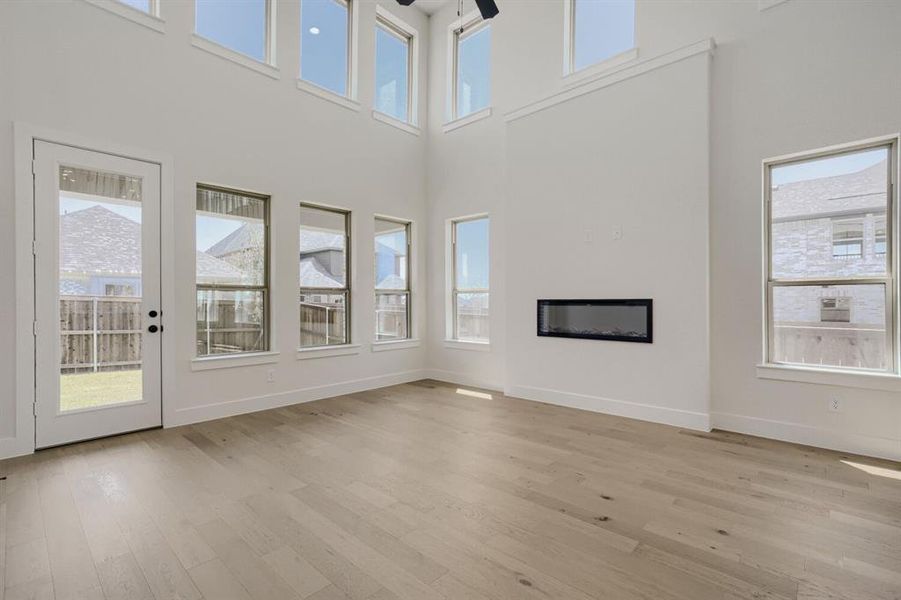 Living area featuring light wood-finish flooring, high ceilings, a wall-mounted linear fireplace, and abundant natural light from multiple windows and a glass-paneled door