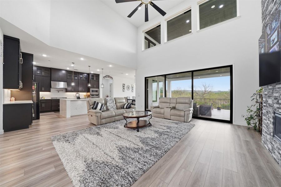 The expansive living space features high ceilings, light wood-style flooring, a floor-to-ceiling fireplace with stone detailing, and large sliding glass doors providing outdoor access