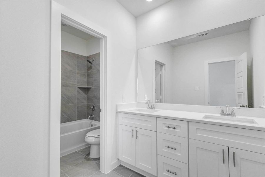 Secondary bath vanity area with dual sinks, white cabinetry, quartz-style countertop, large mirror, and nearby tub/shower room. The layout provides everyday convenience for shared use, while the neutral finishes and clean lines keep the space bright and functional.