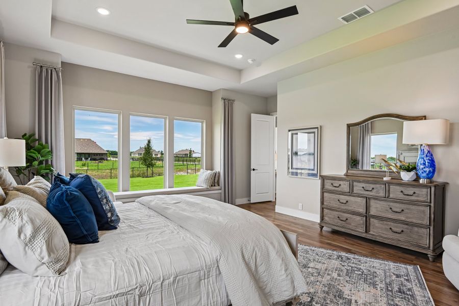 Master bedroom with tray ceiling, ceiling fan, hardwood floors, wood dresser, and large windows with backyard view