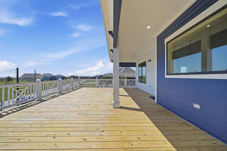 This photo showcases a spacious wooden deck with a white railing, attached to a house with blue siding. The deck offers expansive views of a neighborhood with green lawns and houses, under a bright blue sky. Ideal for outdoor relaxation and entertainment.