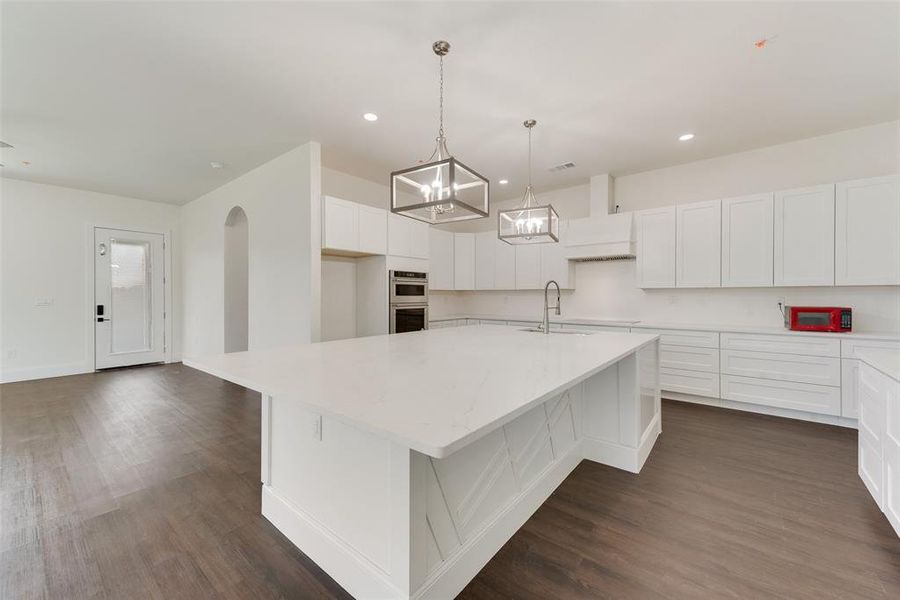 Spacious kitchen featuring a large central island with white countertops, extensive white cabinetry, built-in double ovens, and wood-finish flooring Spacious kitchen featuring a large central island with white countertops, extensive white cabinetry, built-in double ovens, and wood-finish flooring