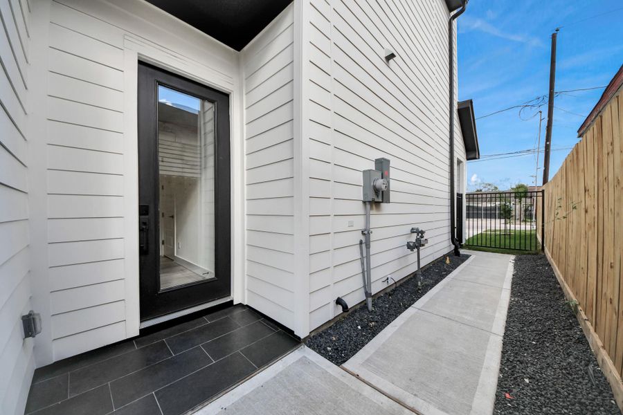 This photo shows a modern side entrance with a sleek black door, white siding, and a narrow concrete pathway. The area is bordered by a wooden fence and features a small, low-maintenance garden with gravel.