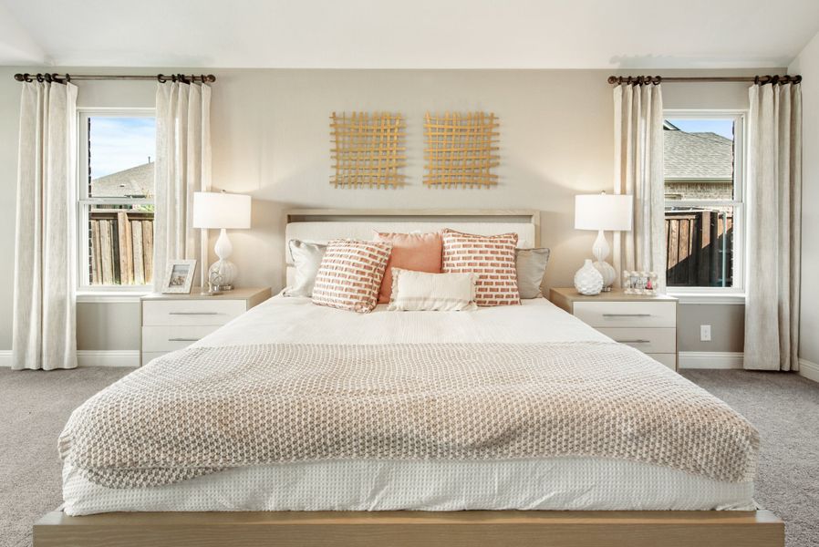 Bedroom with king bed, matching nightstands, two windows with white curtains, and neutral carpet flooring
