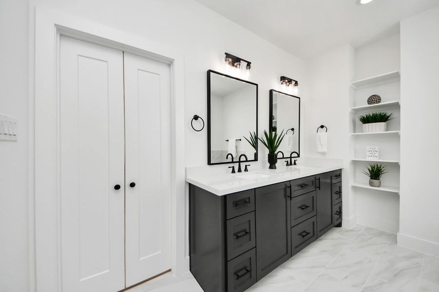 This modern bathroom features a double vanity with sleek black cabinets and large mirrors. It includes elegant fixtures and ample storage, with built-in shelves displaying decorative plants. The space is bright and contemporary, with a clean, sophisticated design.